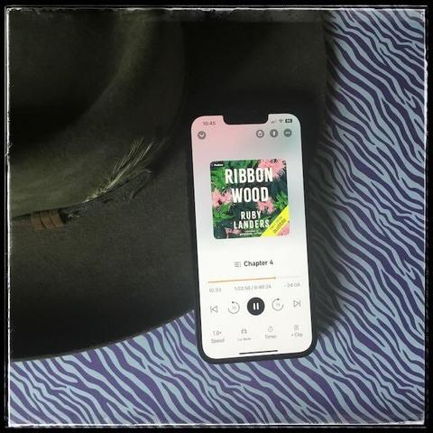 On a purple zebra background, an iPhone playing Ribbonwood by Ruby Landers, narrated by Katherine Littrell, next to an Akubra hat.