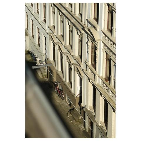 Aus einem Dachfenster heraus blickt man herab auf die gegenüberliegende Fassade und den Bürgersteig. Ein rotes Fahrrad lehnt an einer sandfarbenen und mit Stuck verzierten Hauswand aus der Jahrhundertwende. 