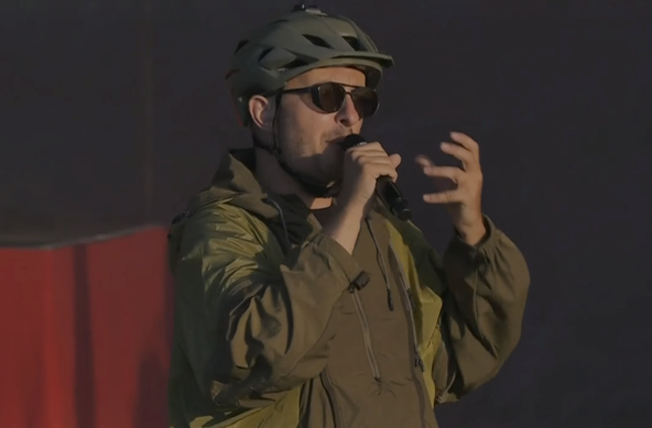 Ein Mann mit Fahrradhelm, Sonnenbrille und Regenponcho in tarngrün hält ein Mikro vors Gesicht.
