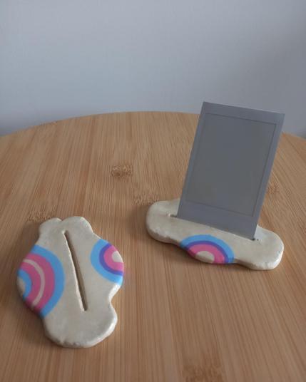 Foto a dos portafotos horizontales con forma de nube. El de la izquierda tiene un arcoiris con los colores de la bandera trans en su lado izquierdo y otro con la bandera bisexual en el lado derecho. El de la derecha tiene una fotografía Polaroid colocada de forma vertical en la ranura central y tiene en su parte delantera pintado un arcoiris con los colores de la bandera bisexual.