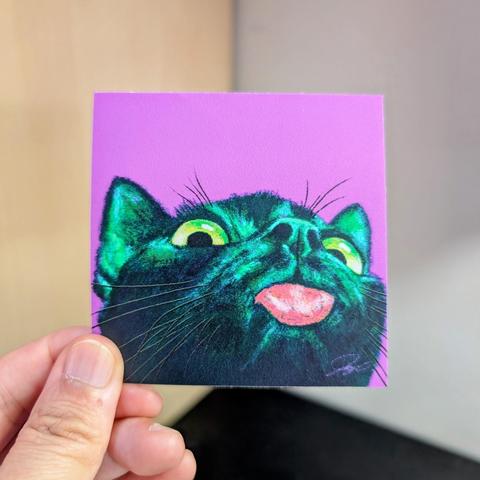 Photo of a left hand holding a square sticker. The sticker is a matte painting of a black cat sticking his tongue out.
