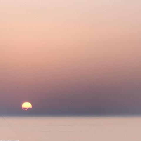 Une photographie de coucher de soleil sur l'océan. Le soleil est près de disparaître derrière la ligne d'horizon. Il est orangé et des nuages masquent sa partie basse. Le ciel est rose-gris.