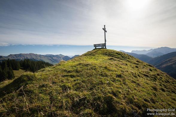 A serene scene captured on a hill, showcasing a wooden bench with a simple cross on top. The hill overlooks a vast landscape with a clear blue sky and fluffy white clouds. The setting is peaceful and tranquil, surrounded by mountains and greenery. The dominant colors in the image are white and a warm brown accent. The image exudes a sense of spirituality and contemplation, inviting viewers to sit and reflect on the beauty of nature. This picturesque hill station offers a perfect spot for relaxa…