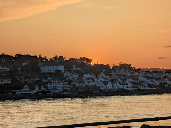 The white wooden houses of outer Langesund, seen from across the sound, with the sun sliding behind the hill top. The sound with its silvery sea lights up the foreground.