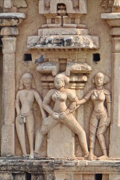 Sculpture group from Virupaksha Temple, a temple dedicated to Lord Shiva that dates to the 7th century. The temple is located on the banks of the Tungabhadra River in Hampi, India. The scene depicts a nude woman standing with her legs spread widely. She spreads her vulva with both hands. To her right stands a man with a semi-erect phallus who touches her thigh. To her left, a woman wearing a belt-like garment or jewellery touches her arm.