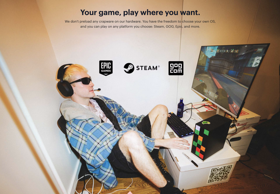 A person with blonde hair and sunglasses wearing headphones with a microphone, sitting in front of a computer monitor displaying a game. They are wearing a blue and white plaid shirt and dark shorts. On the desk are a keyboard, a mouse, and a black Framework Desktop computer tower with colored squares on the side, standing upright. Logos for Epic Games, Steam, and GOG.com are visible above the person.