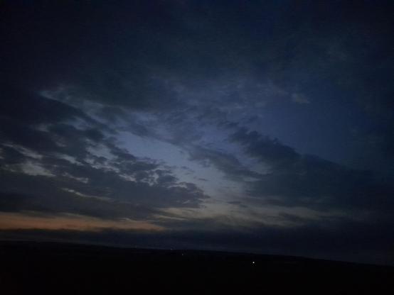 Dark flat landscape dotted with a few distant specks of lights, under a midnight blue sky too light for the stars to be visible, with streaks of orange-red and light blue, partially obscurer by grey clouds with silvery edges.