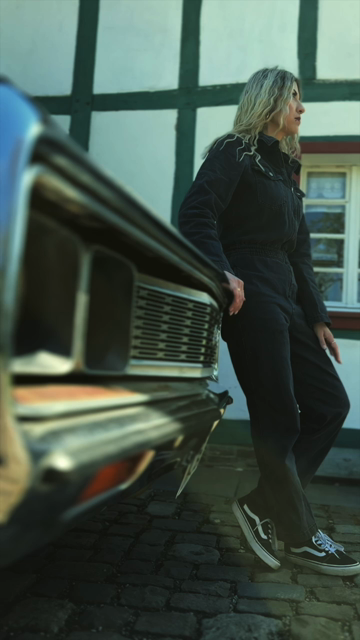 Das Video beginnt mit einer Person, die an einem klassischen Auto lehnt, während sie in einem traditionellen Fachwerkhaus steht. Sie trägt eine schwarze Jeansjacke und hat blonde, wellige Haare. Der Hintergrund zeigt ein weißes Fachwerkhaus mit grünen Balken. Die Person steht dann vor dem Haus, und auf dem Bildschirm erscheint der Text "Hey, hier ist Kim von DARKK!" in gelber Schrift. Der Text wechselt zu "Du hast unsere 1. Single HOME geliebt?" und schließlich zu "Mach Dich bereit für unsere n…