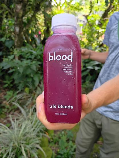 Someone holding out a bottle of juice named "Blood" there are a lot of plants in the background. The juice includes beets as an ingredient so it is quite red, but not the color of blood particularly.