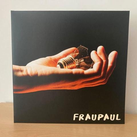 Ein Albumcover mit einer Hand, die eine kaputte Glühbirne hält. Der Hintergrund ist schwarz, und der Name des Künstlers "FRAUPAUL" ist in fetten weißen Buchstaben am unteren Rand zu lesen.