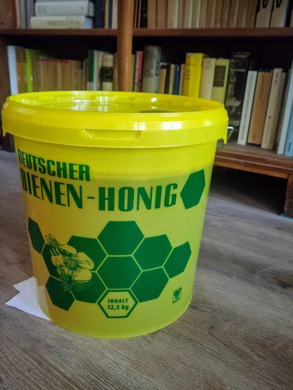 Ein großer Eimer, gelb und halbtransparent, fast ganz gefüllt, mit dem Aufdruck "Deutscher Bienen-Honig" steht vor einem Bücherregal.