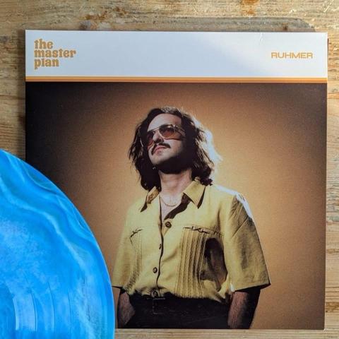 Ein Albumcover, das einen Mann mit Sonnenbrille und Schnurrbart zeigt, der ein gelbes Hemd trägt. Der Hintergrund hat einen warmen Ton, und eine blaue Schallplatte ist teilweise im Vordergrund zu sehen. Der Text auf dem Cover lautet "the master plan".