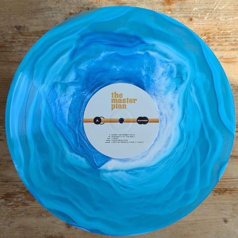 Eine leuchtend blaue Vinyl-Schallplatte mit dem Titel "The Master Plan", die ein wirbelndes ozeanähnliches Design aufweist. Auf dem Etikett in der Mitte stehen Titel wie "Down the Rabbit Hole" und "Light My World (Take It Easy)".