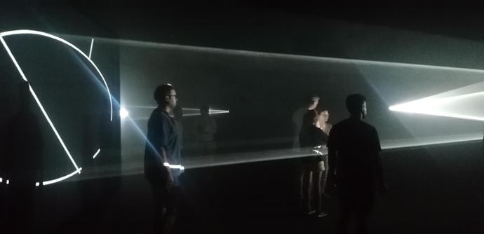Lichtstrahlen bilden geometrische Formen und ein paar Menschen laufen vorbei im Dunkeln - nur die Strahlen und Schemen sind sichtbar