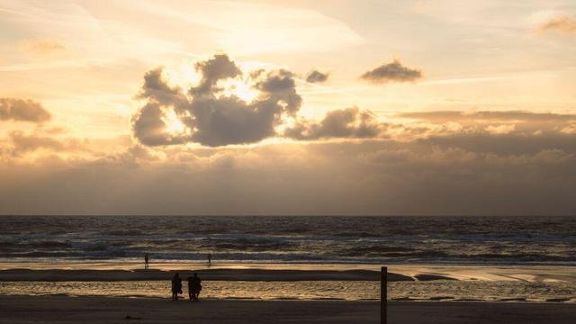 Zonsondergang aan het strand bij De Koog op Texel. Er zijn wat wolken aan de hemel, waarvan een de zon afschermt en waardoor er wat Jakobsladders te zien zijn. De sfeer is warm en geel.  Aan de vloedlijn zijn wat wandelaars te zien.