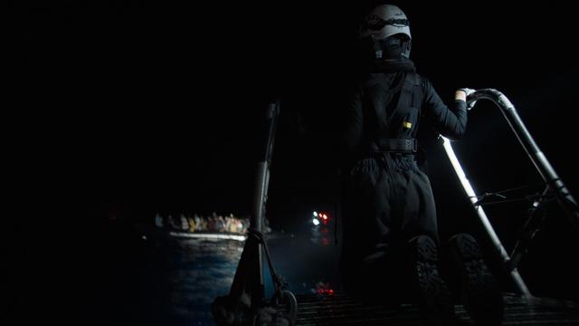 Es ist noch dunkel. Auf einem RHIB steht ein*e Seenotretter*in und blickt in Richtung eines völlig überfüllten Bootes, das im Scheinwerferlicht nur unscharf auszumachen ist.