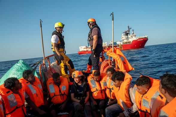Mehrere Menschen mit orangen Schwimmwesten sitzen in einem kleinen Boot auf dem Meer, zwei Rettungskräfte
