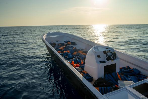 Kleines Boot auf offenem Meer bei Sonnenuntergang, gefüllt mit vielen blauen und orangefarbenen Rettungswesten.