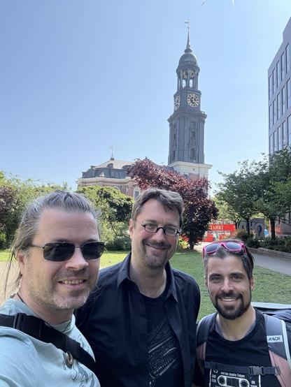 Drei Männer stehen lächelnd im Vordergrund eines sonnigen Außenfotos. Sie posieren für ein Selfie in einem kleinen Park. Alle tragen Freizeitkleidung und Rucksäcke, zwei von ihnen eine Sonnenbrille. Im Hintergrund ist die barocke Turmfassade der St. Michaelis Kirche („Michel“) in Hamburg zu sehen. Der Himmel ist klar und blau, umgeben von grünen Bäumen und einem modernen Bürogebäude am rechten Bildrand.
