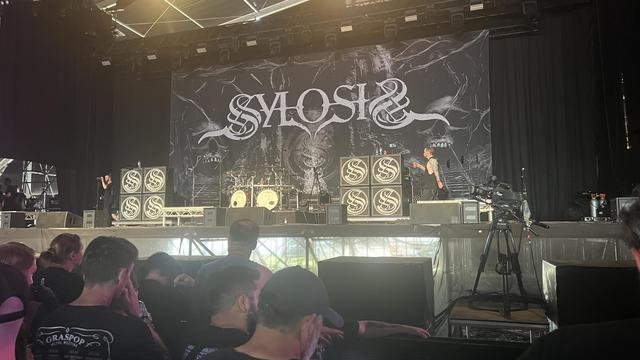 Das Bild zeigt eine Live-Auftrittsszene einer Band namens "SYLOSIS" auf einer Bühne. Im Hintergrund ist ein großes Banner mit dem Bandlogo zu sehen, das in einem düsteren, gotischen Stil gestaltet ist. Auf der Bühne stehen mehrere große Lautsprecher mit dem Bandlogo, und es sind Musiker zu sehen, die sich auf der Bühne befinden. Einige Musiker sind mit Instrumenten wie Gitarren und Schlagzeugen zu sehen. Vor der Bühne sitzen und stehen Zuschauer, die das Konzert verfolgen. Einige Zuschauer tragen schwarze Kleidung, was typisch für Metal-Konzerte ist. Ein Kameraschwenkertisch ist auf der rechten Seite der Bühne zu sehen, was darauf hinweist, dass das Konzert aufgezeichnet wird. Die Atmosphäre wirkt energiegeladen und intensiv, was für ein Metal-Konzert typisch ist.

Bereitgestellt von @altbot, privat und lokal generiert mit Ovis2-8B

🌱 Energieverbrauch: 0.230 Wh