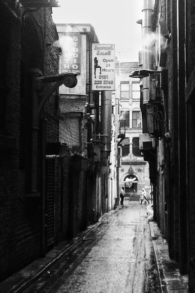 Black and white photo of a narrow street with a sign on the top left and some people walking at the end of the street.