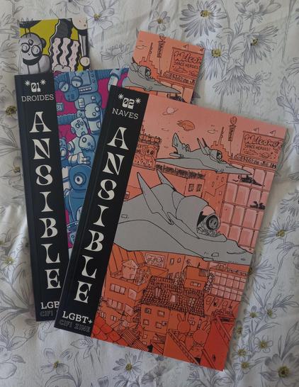 Foto a los dos primeros números del fanzine Ansible de Bronco Cómics sobre una colcha de flores.
El primer número se titula "Droides" y el segundo, "Naves". En ambos se lee el número, el título, "Ansible" y "LGTB+ cifi zine" en la parte izquierda de cada tomo en texto blanco sobre fondo negro. En el resto de sendas portadas, hay una ilustración que representa el tema (robots bailando en el 1, naves sobrevolando una ciudad en el 2). Sendos tomos van acompañados de un marcapáginas que sobresale de cada uno.