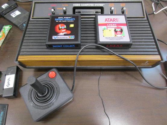 Atari VCS console with a joystick out front and two ROM cartridges on top, "Secret Collect" and "C.H.E.A.T."
