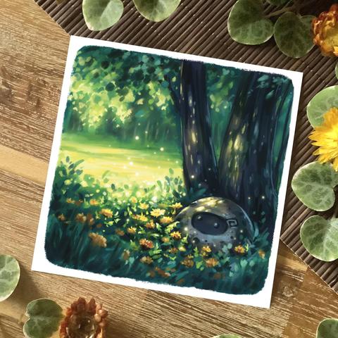 A photo of a square postcard placed on a wooden table and a mat, surrounded by yellow flowers and leaves for decoration. The postcard features a digital painting of a little rocky spirit sitting in the half-shade under a tree, surrounded by dandelions. A meadow flooded with sunlight and a treeline are visible indistinctly in the background.