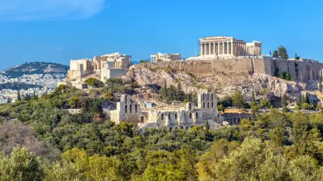 Photograph of the Athenian acropolis on a clear summer day. The sky is blue and the trees are a lush green, the modern buildings only visible in the far distance with the ancient acropolis and theatres in focus.