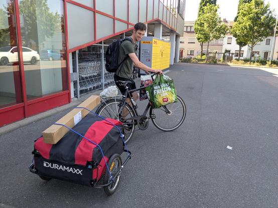 Man stapt op een fiets, waar aan het stuur 2 grote boodschappentassen vol boodschappen hangen. Achter de fiets een brede kar, ook vol boodschappen. Afgedekt met een rood zwart zeiltje. En dat bovenop nog een langwerpig kartonnen pakket ( parasol??). Hij glimlach en zei vol vertrouwen dat hij goed thuis ging komen met de spullen voor het verjaardagsfeestje.