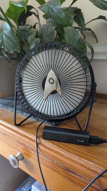 A photo of a USB-powered fan, sitting on a table, plugged into to a small portable power bank. The fan has a magnetic, silver Star Trek comm badge attached to the centre of the casing.