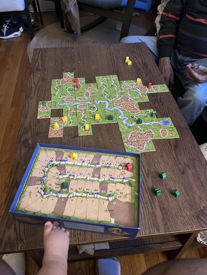 A tabletop game scene featuring a Carcassonne board with several tiles depicting roads, cities, and fields. Colorful wooden meeples in yellow, red, green, and pink are placed on the board. Nearby, there is a secondary board game score card. In the very bottom of the photo there is the foot of an infant on a lap.