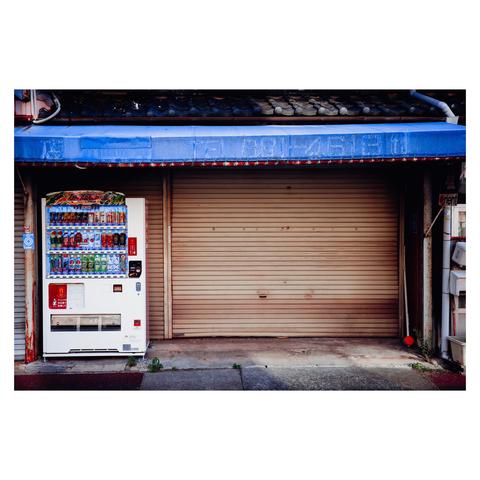 The image shows a vending machine standing alone in front of a closed storefront in Nagoya. The metal shutter is beige and weathered, with visible rust and stains. Above it, a blue awning with faded lettering hangs loosely, remnants of a store that’s either on break or long since retired. To the left, the bright white vending machine is fully stocked with rows of colorful drinks, including sodas and canned coffee.