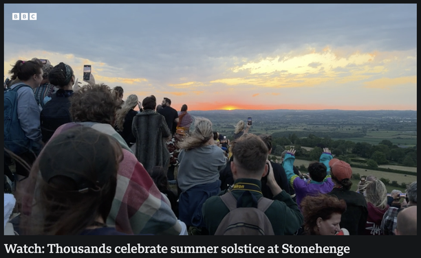 The image depicts a large group of people gathered on a hilltop, facing the horizon where the sun is setting. The sky is painted with hues of orange, pink, and blue, indicating the time is around sunset. The crowd is diverse, with individuals of various ages and attire, some wearing hats and others with backpacks. Many people are using their smartphones to capture the moment, with their arms raised towards the sky. The landscape in the background features rolling hills and fields, typical of th…