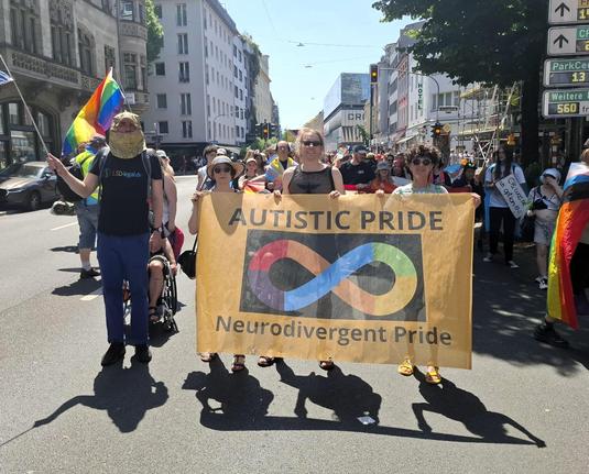 Foto zeigt eine Fußgruppe mit dem goldenen Autistic Pride Banner. Darauf sind das regenbogenfarbene Unendlichkeitszeichen und die Schriftzüge Autistic Pride und Neurodivergent Pride abgebildet.