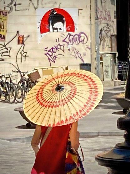 On a Barcelona street, a person wearing a red dress holding an Asian umbrella with red accents walks through a street with graffiti-covered walls. In the background, there is a red and white art piece depicting a face. Bicycles are parked nearby. #SilentSunday 20250622 © Gilles Denizot