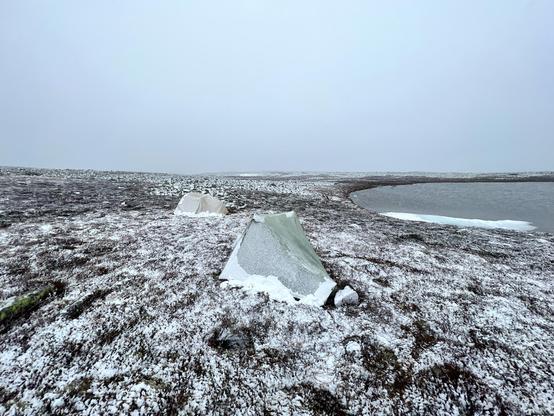 Two snow covered tents (just very little) next to a lake. Barren landscape, no trees. 