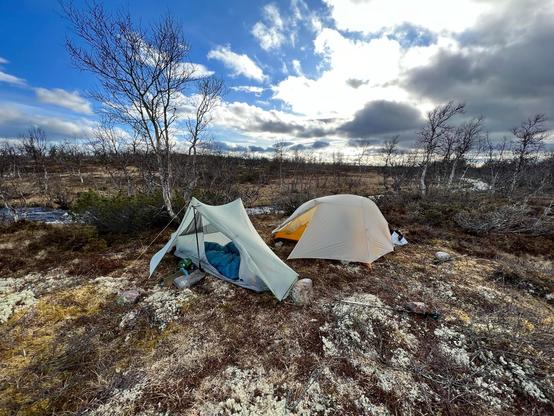 Two tents between sparse birch trees on otherwise barren landscape. There’s a small stream behind the tents. Cloudy sky with patches of blue. 