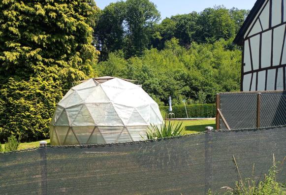 A geodesic dome as a greenhouse in a garden. In the background trees, to the right a wall of a half-timbered house.