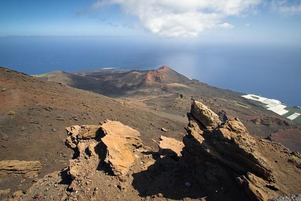 Fotografía en la q se ve en primer plano tercio inferior unas rocas volcánicas en el centro un cono volcanico, en la parte superior el cielo y el mar y en el lado derecho unas salinas en la costa.