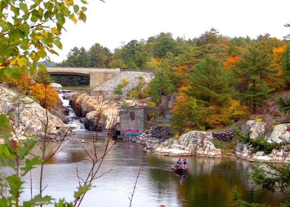 Waterfall under a roadway with river leading away. Boat with two occupants fishing. Autumn scene.