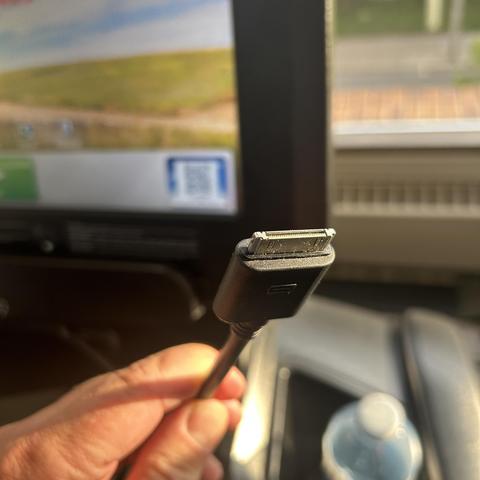 An individual is holding a black cable with a rectangular connector that is an old wide Apple connector for early iPods- against a blurred background featuring a screen and a water bottle.