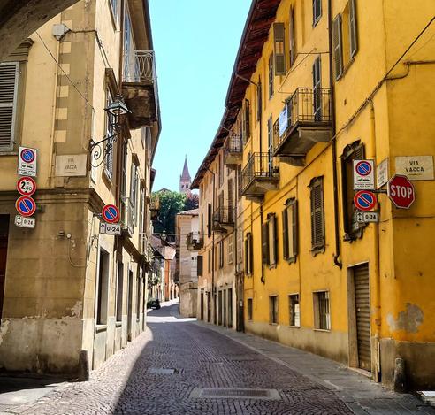 Blick durch eine schmale Gasse in einer italienischen Stadt. Das rechte Haus ist kräftig gelb, die Straße isz gepflastert. Im Hintergrund ist ein Kirchturm zu sehen. 