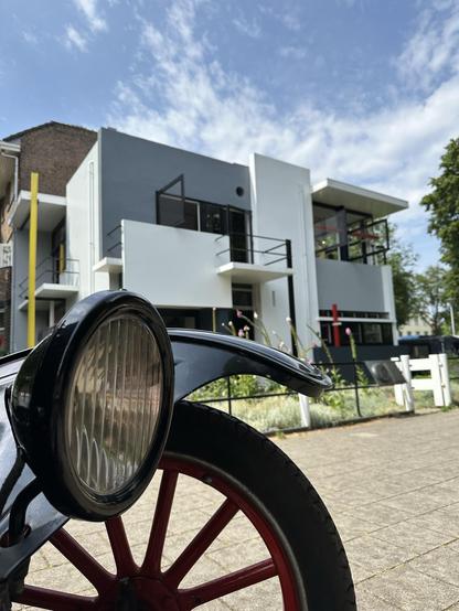A round headlight from a Ford T in the foreground and a modern white and grey building with black, yellow and red accents in the background: the Rietveld Schröder House in Utrecht.