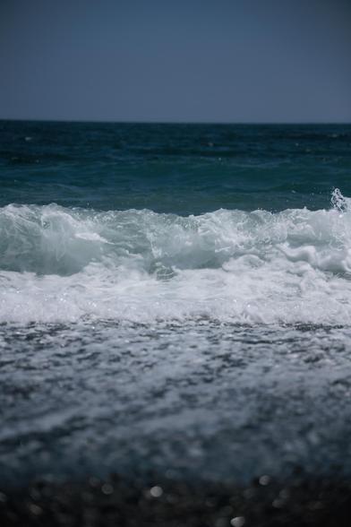 Waves crashing onto a pebbly shore, creating white foam against a backdrop of deep blue ocean under a clear sky.