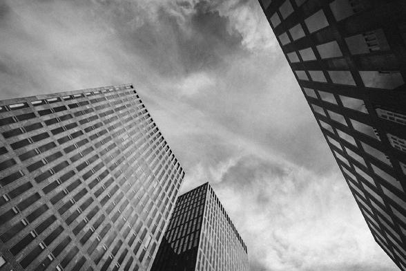 Dramatic black and white photo looking up at three modern high-rise buildings with geometric window patterns, set against a cloudy sky with strong contrast and texture.
