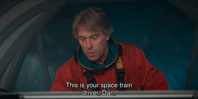 Jodie Whittaker episode. Dan is wearing one of those New Doctor Who orange space suits that’s actually designed for the surface of Mars and not outer space, and as such, is made of a thinner material that doesn’t need as much pressurization against the vacuum of space, but nonetheless, the prop is being reused as a space suit, which he is wearing without his helmet because he inside a generic dark smoky room with arching gray walls, as though they did not have the budget to build a real set, looking down at a screen. Caption reads, “This is your space train driver, Dan,”