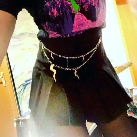 A close-up of a person wearing a black skirt and a graphic t-shirt featuring vibrant colors. The attire includes a silver chain belt adorned with lightning bolt charms. The background features a wooden door and a blurred painting.
