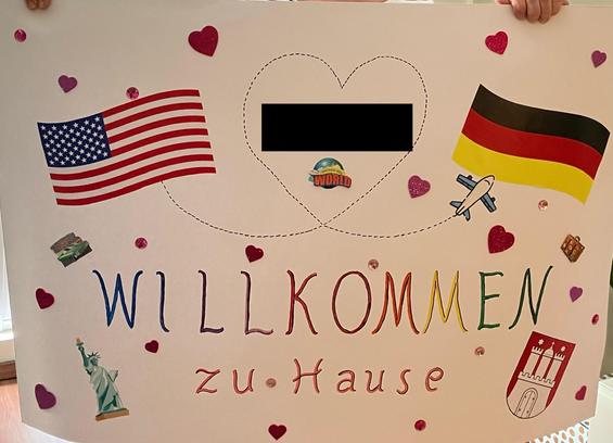 Plakat Willkommen zu Hause mit USA und Deutschland Flagge Freiheitsstatue und dem Hamburg Wappen. Ein Flugzeug fliegt ein Herz in dem ein geschwärzter Name steht.