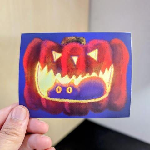 Photo of a left hand holding a rectangular sticker. The sticker is a matte drawing of a black cat in a glowing jack o lantern.
